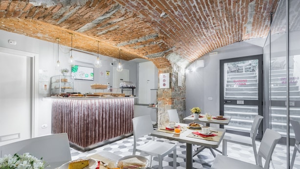 A breakfast area with a brick ceiling, white walls, and a tiled floor. There are tables with breakfast items, chairs, and a counter with a display case. - Hotel Cosmos (Florence)