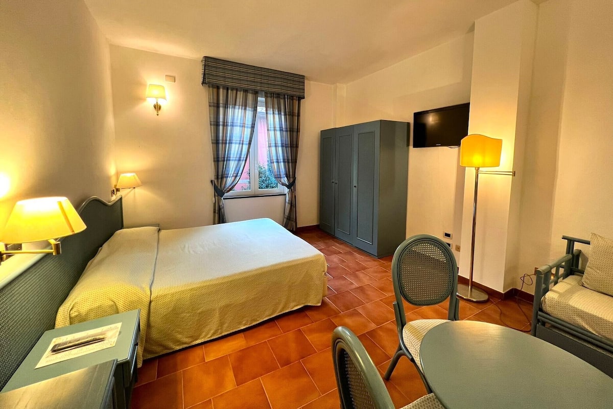 Ein Hotelzimmer mit zwei Betten, einem Kleiderschrank, einem Schreibtisch mit Lampe, einem Sessel und einem an der Wand befestigten Fernseher.