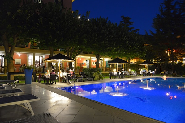 A poolside area with lounge chairs, a dining area, and a building with illuminated windows.