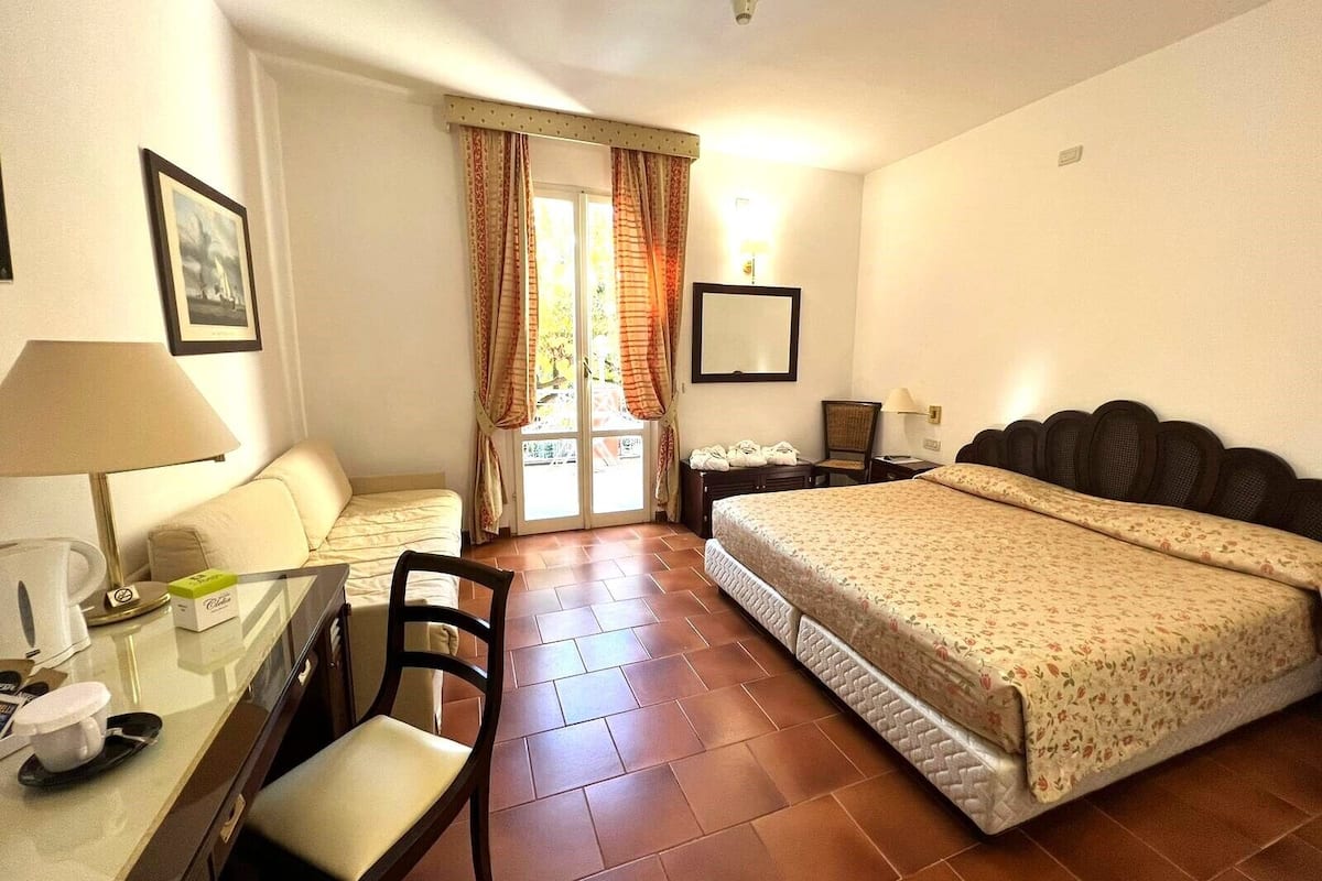 Ein Hotelzimmer mit Bett, Schreibtisch mit Stuhl, einem kleinen Tisch mit Lampe, einem Sessel sowie einem Fenster mit Vorhängen und einem Wandspiegel.