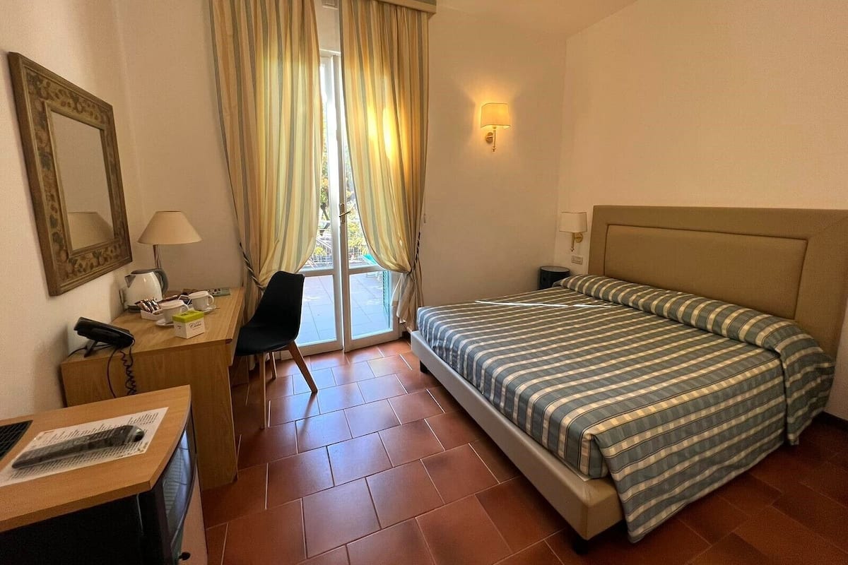 Ein Hotelzimmer mit Bett, Schreibtisch mit Telefon, Stuhl, Lampe und einem fensterbepflanzten Fenster.