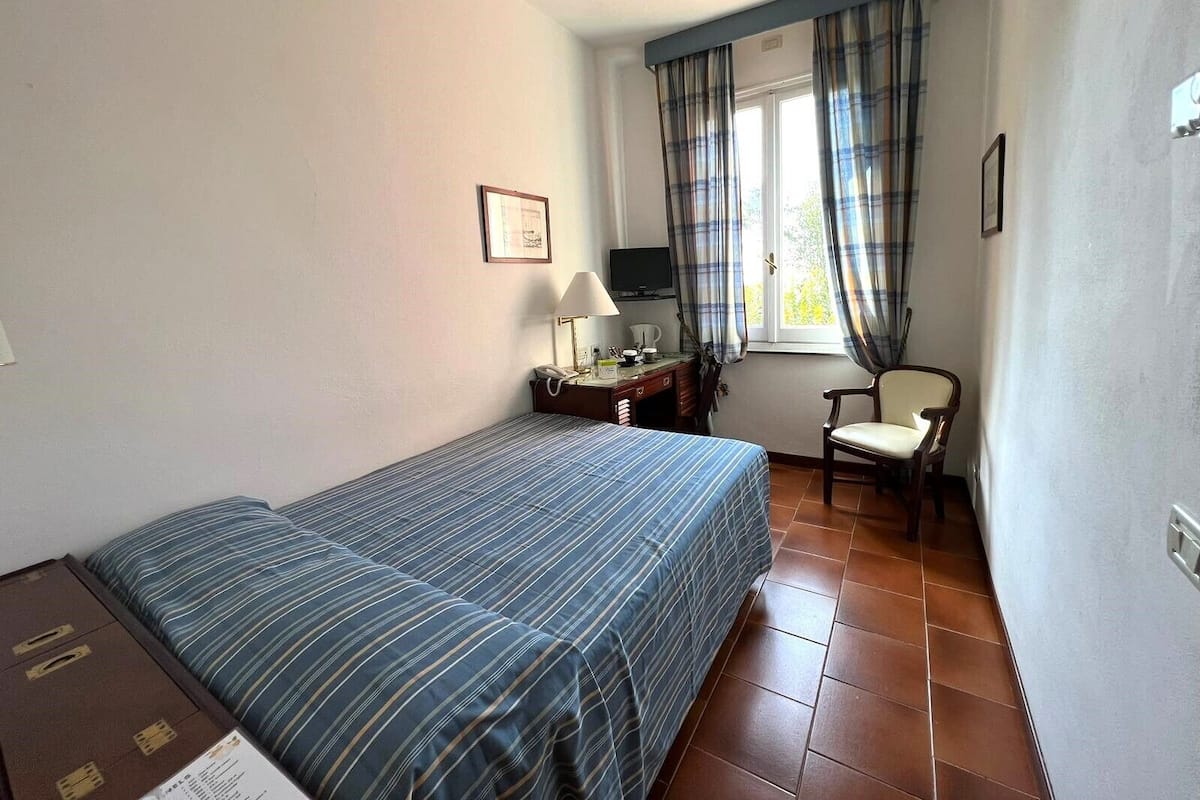 Ein kleiner, ordentlicher Raum mit einem Bett, einem Schreibtisch mit Stuhl, einem Fernseher und einem Fenster mit Vorhängen.