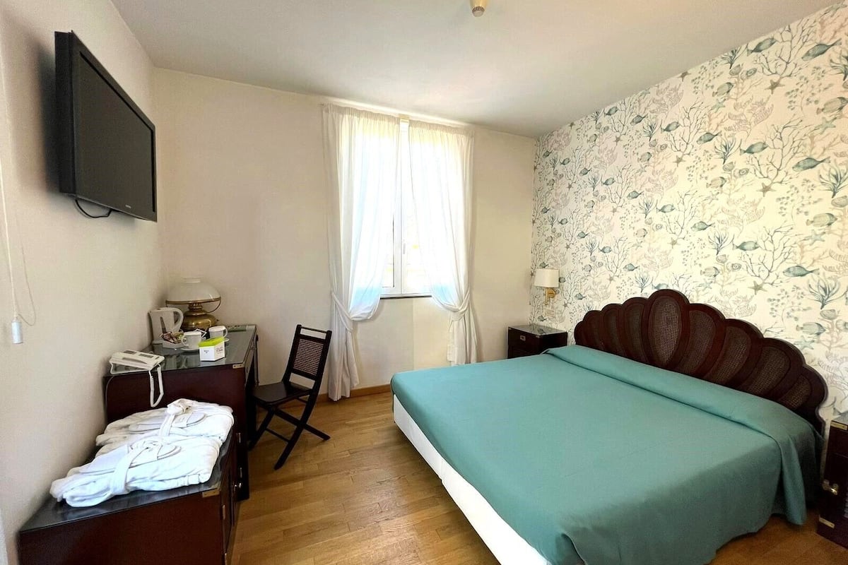 Ein Hotelzimmer mit Bett, Schreibtisch mit Telefon, Stuhl, Fenster mit Vorhängen und einer Wand mit Blumenmuster.