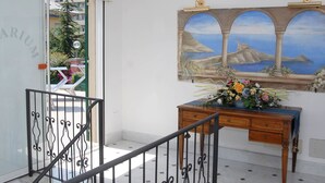 Um quarto com porta de vidro, escada com grades de ferro forjado, escrivaninha de madeira com arranjo floral e um mural de paisagem costeira.