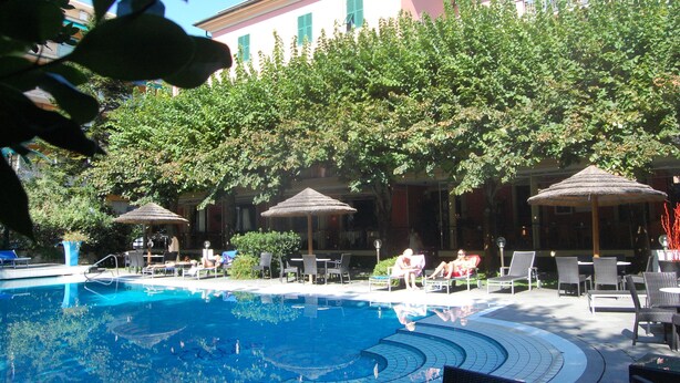 Área de piscina com espreguiçadeiras e guarda-sóis, cercada por árvores e um edifício ao fundo.