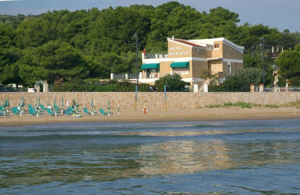 Un hôtel appelé «Hotel Velia Velo», avec vue sur la plage, un mur en pierre et des fauteuils de salon verts.