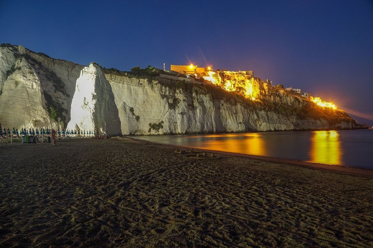 A coastal cliff with a building illuminated at night, a sandy beach, and calm water reflecting the lights.