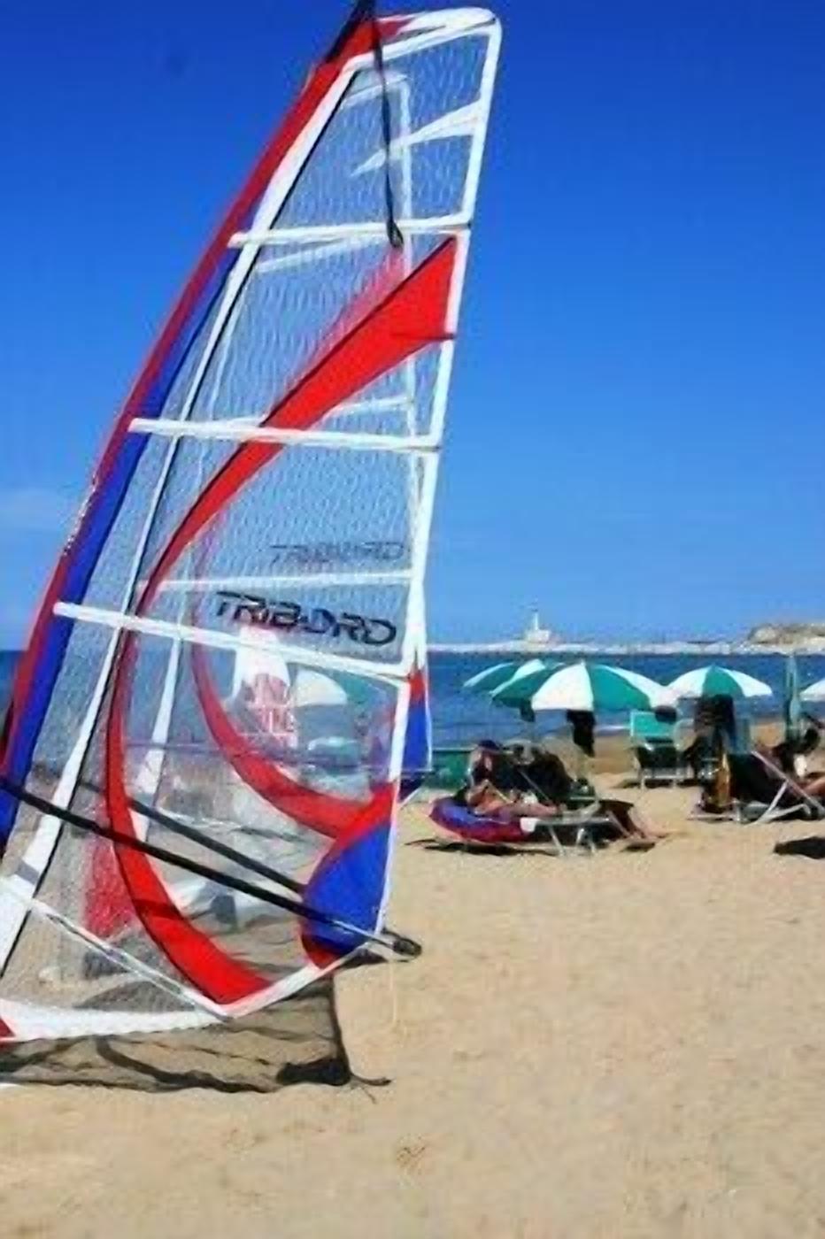 Tabla de windsurf con un diseño rojo, blanco y azul en una playa con sombrillas y gente al fondo.