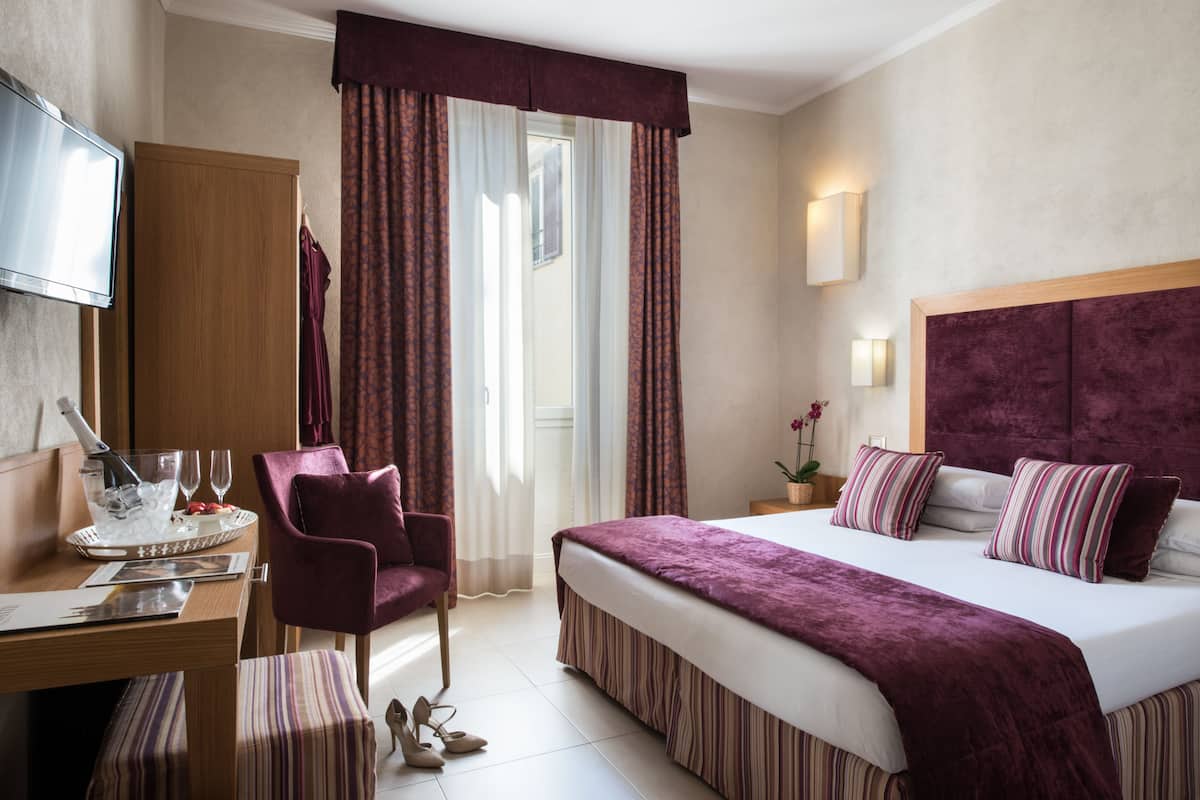 Une chambre d’hôtel avec un lit, un bureau et une chaise, une télévision et une fenêtre avec des rideaux.