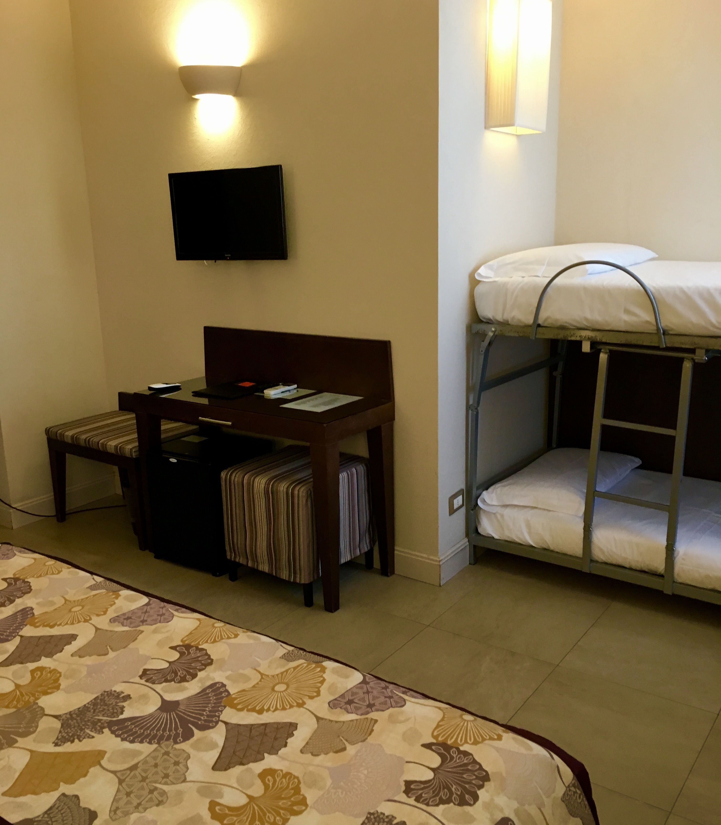 A hotel room with a bed, a desk, a chair, a bunk bed, and a flat-screen TV mounted on the wall.