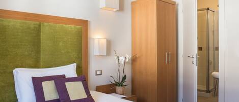A hotel room with a bed, a wooden wardrobe, a nightstand, a wall-mounted light, and a potted orchid.