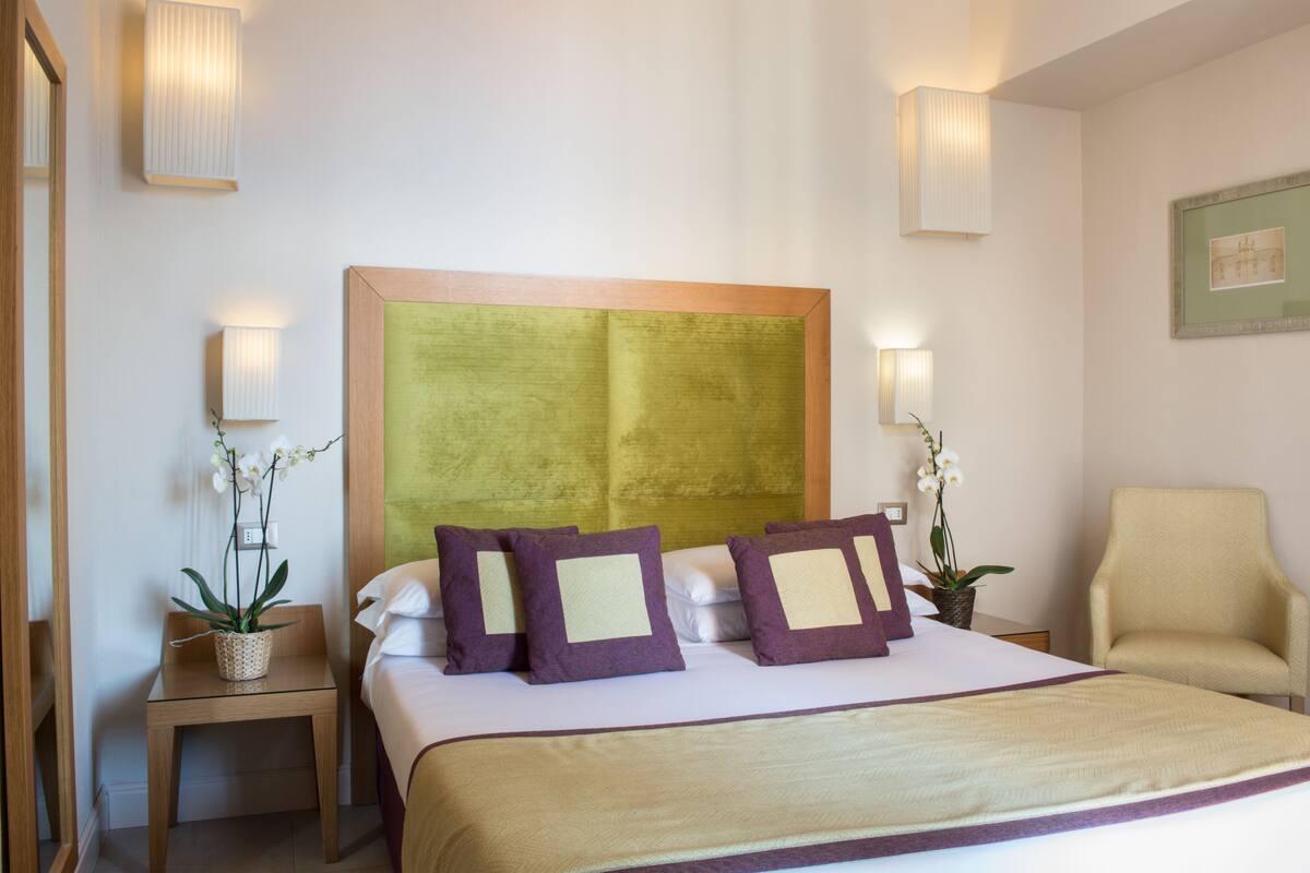 Une chambre d’hôtel avec un grand lit, deux tables de chevet, une chaise et des lampes fixées au mur.