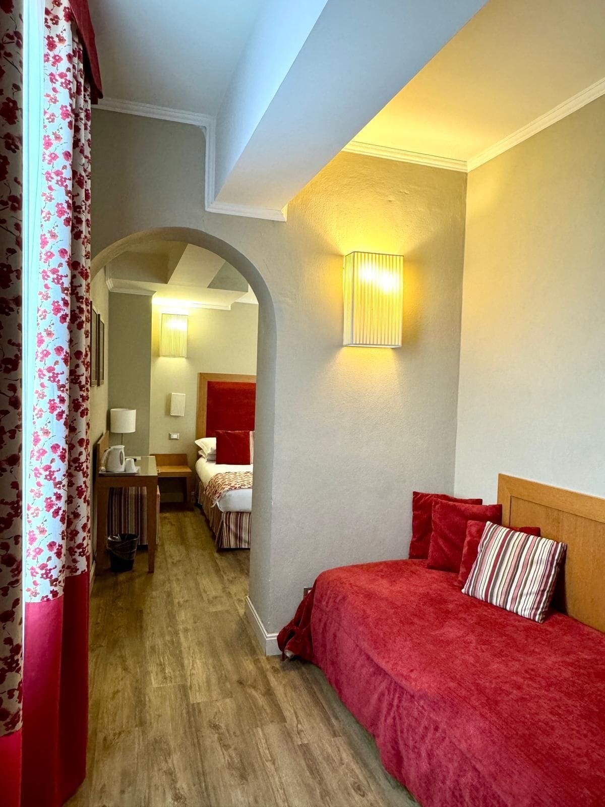 A hotel room with a wooden floor, a bed with red bedding, a nightstand with a lamp, and a wall-mounted light fixture.
