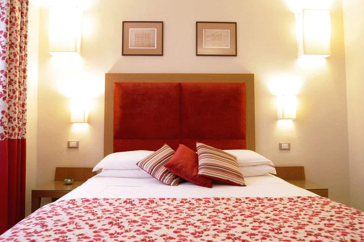 Une chambre d’hôtel avec un grand lit, une tête de lit rouge, deux tableaux encadrés au mur et deux lampes de chevet.
