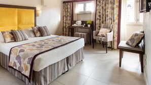 Habitación de hotel con una cama grande, una mesita de noche con una planta, una silla, un escritorio con lámpara y una ventana con cortinas.