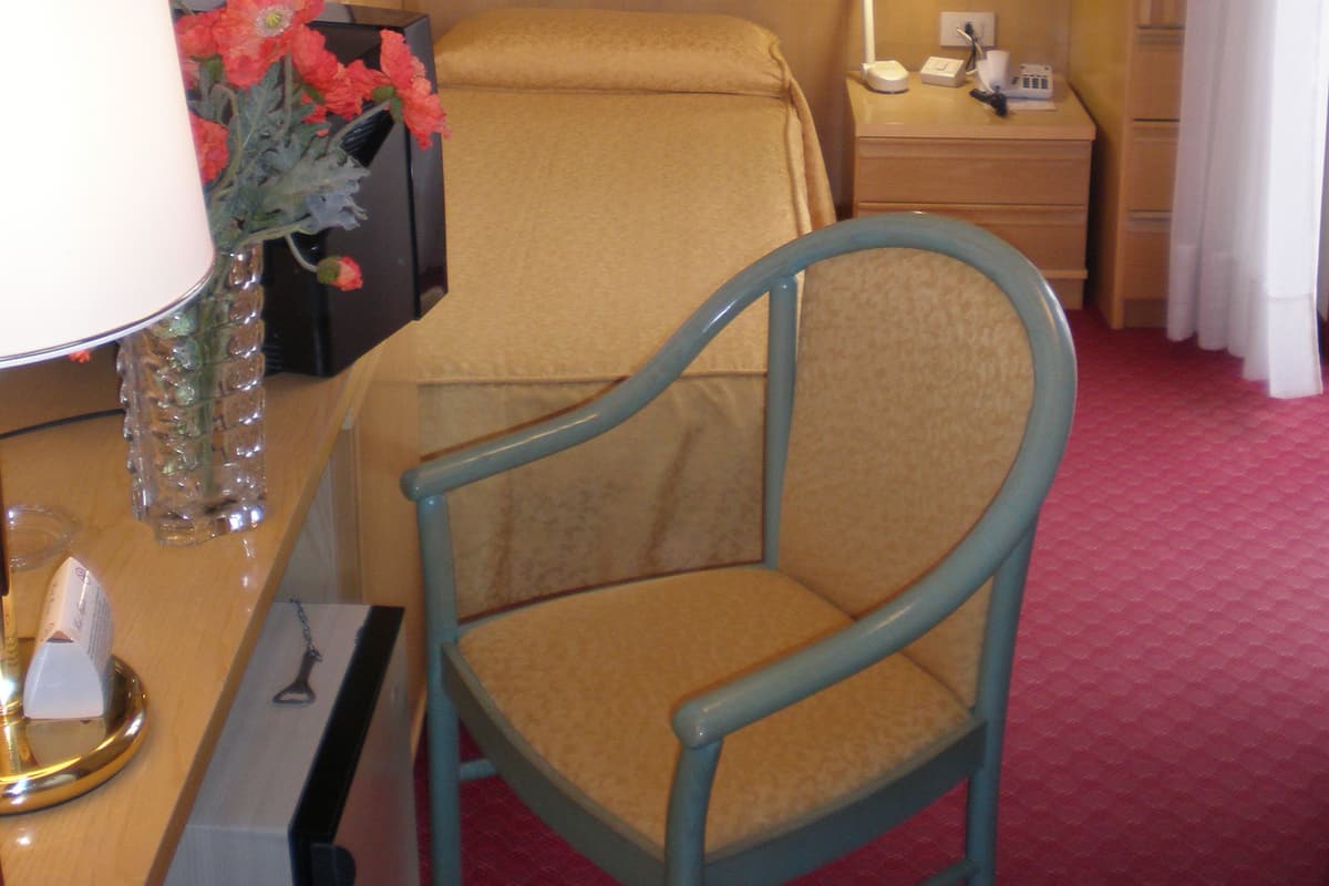 A hotel room with a wooden desk, a chair, a lamp, a vase with flowers, and a bed.