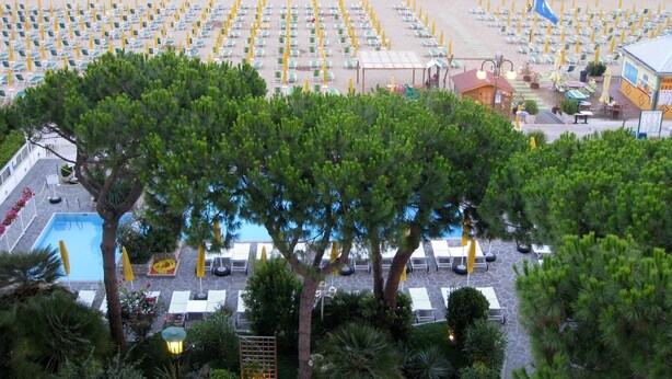 A beachside resort with rows of sun loungers and umbrellas, a pool area, and a building with a sign.