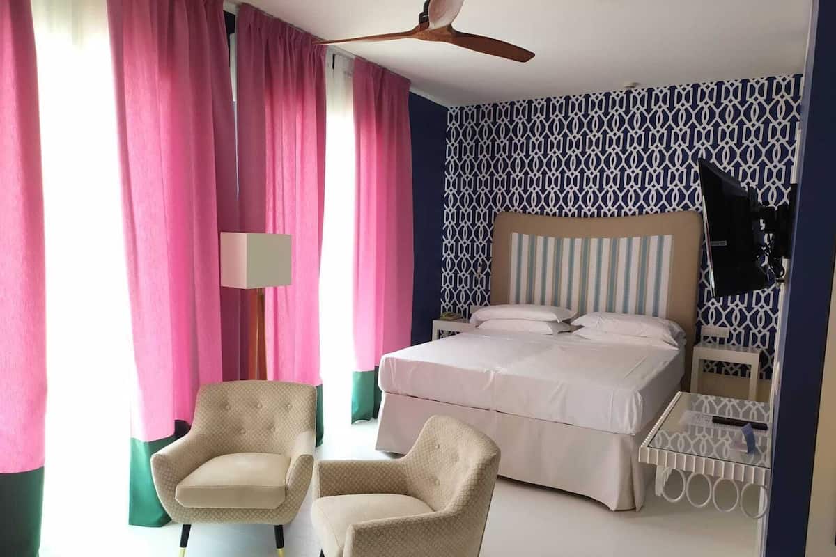 A bedroom with a bed, two armchairs, a ceiling fan, a bedside table, and a patterned wall.