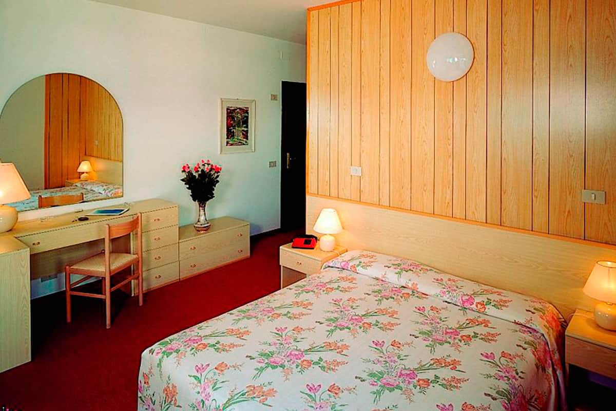 A bedroom with a bed, a desk, a chair, a dresser, a mirror, and a wall-mounted lamp.