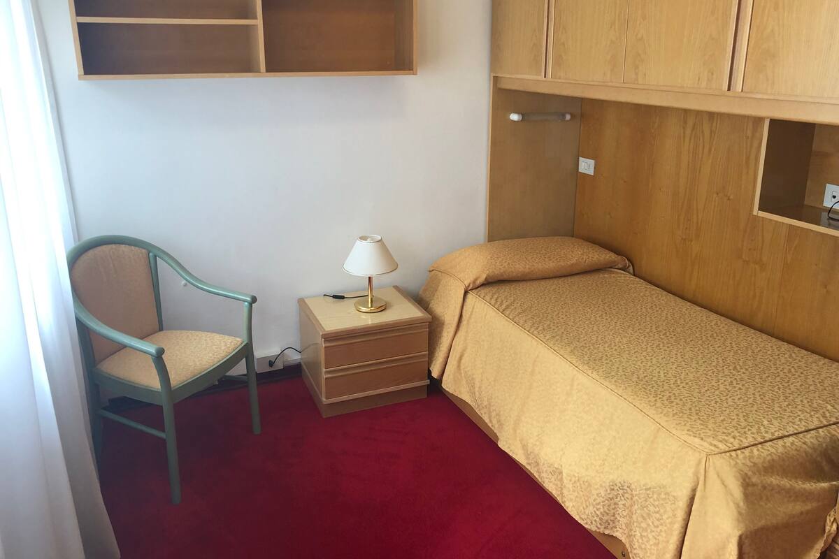 A small room with a single bed, a chair, a nightstand with a lamp, and a wooden shelf above the bed.