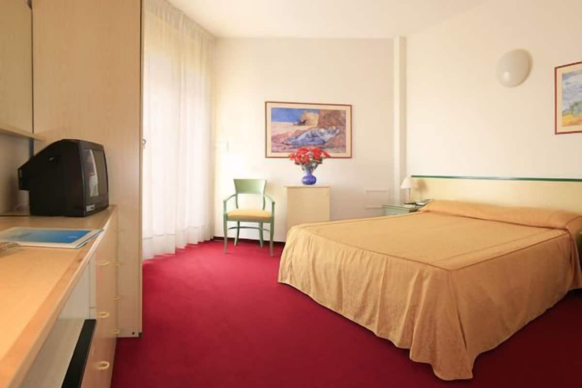 A hotel room with a bed, a desk with a television, a chair, a vase with flowers, and a painting on the wall.