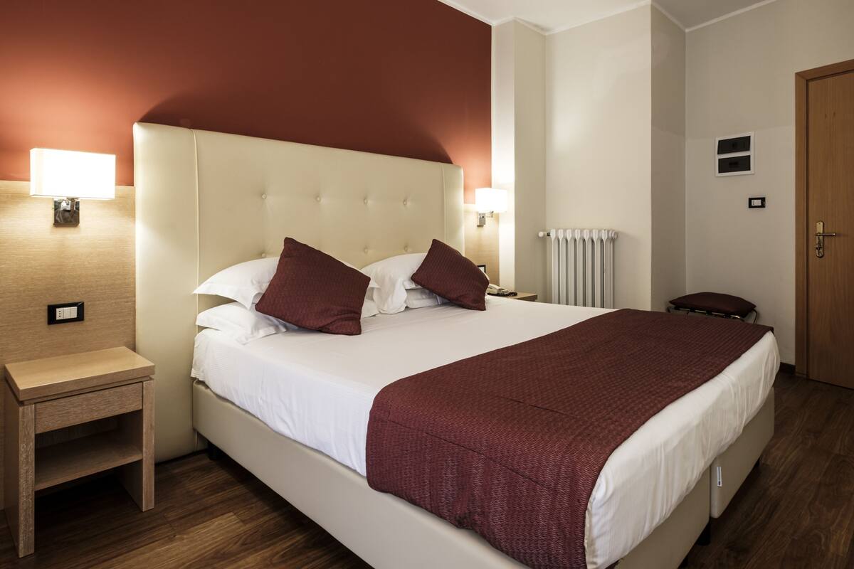 A hotel room with a large bed, two bedside lamps, a wooden nightstand, a radiator, and a door.