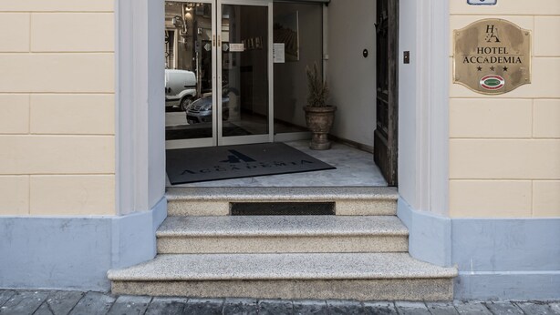 Entrada de un hotel con un letrero que dice "HOTEL ACCADEMIA" sobre la puerta, una planta en maceta y una placa con el número "6".