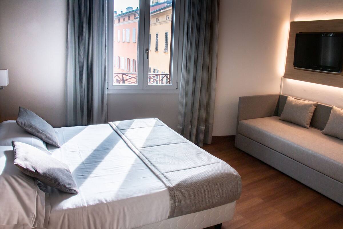 A hotel room with a bed, a sofa, a TV mounted on the wall, and a window with curtains.