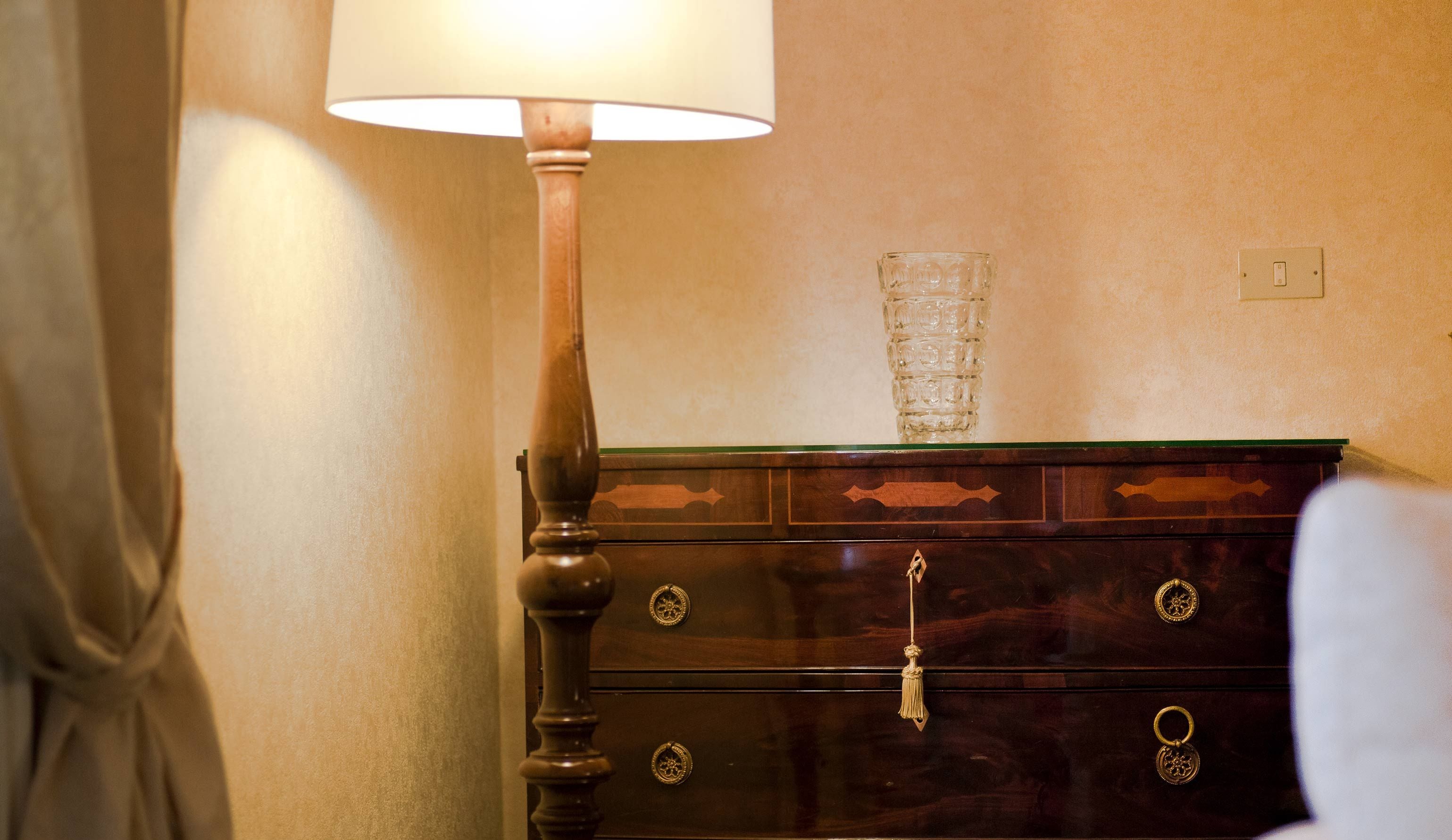 A wooden dresser with a glass vase, a lamp, and a light switch on the wall.