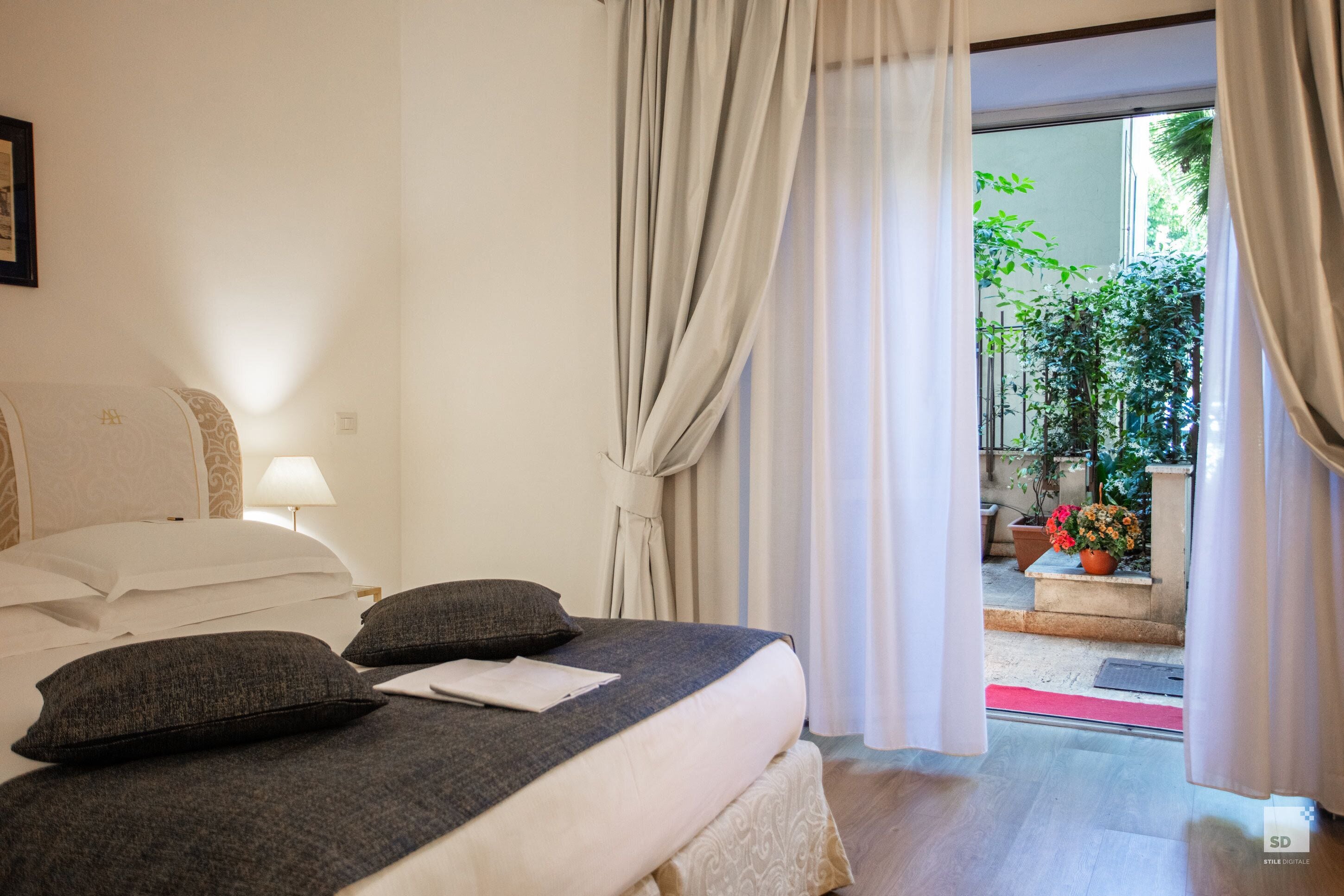 A hotel room with a bed, pillows, a nightstand, a lamp, and a view of the outdoor garden through the curtains.