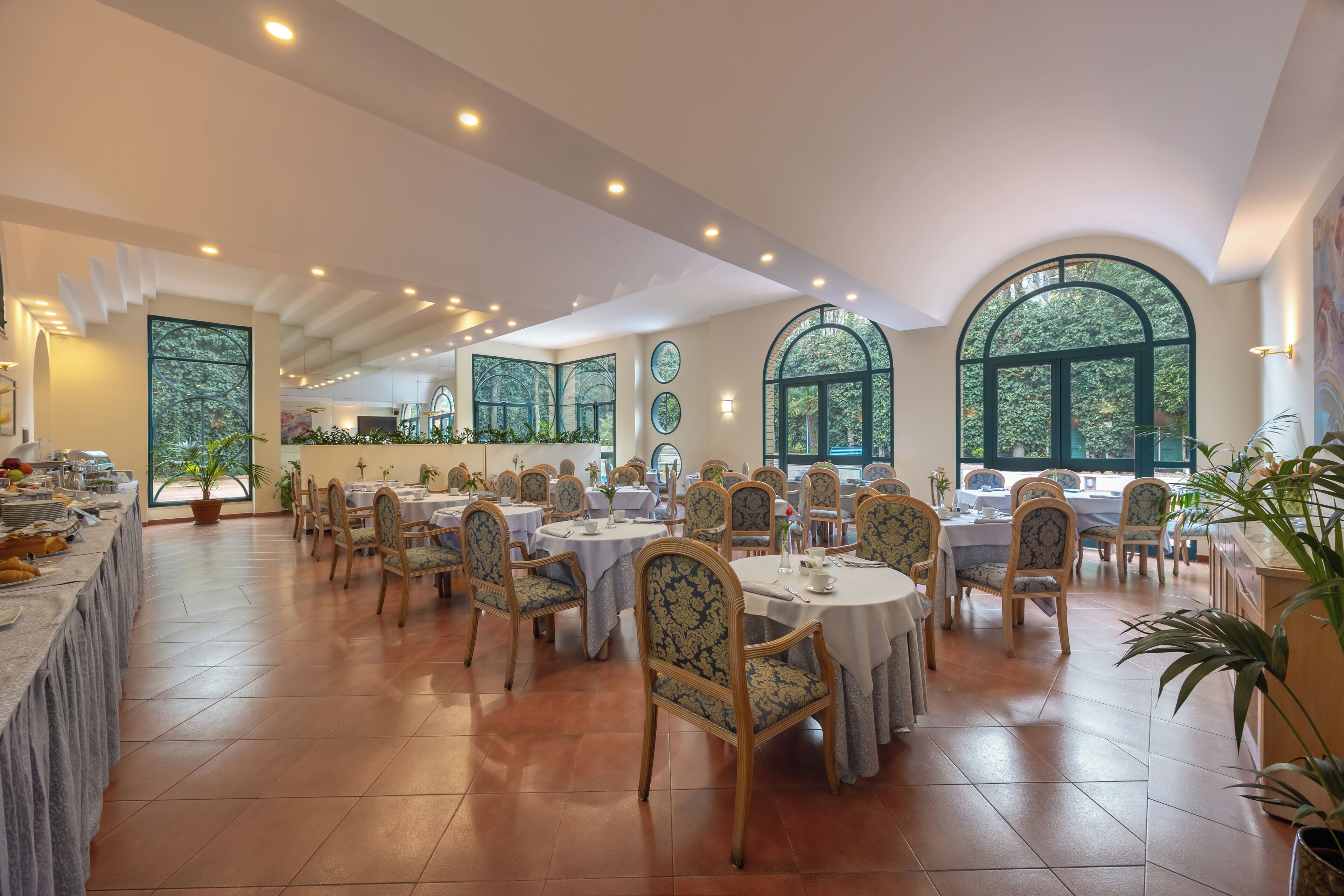 Un'ampia sala da pranzo con tavoli e sedie rotondi, grandi finestre ad arco e un'area buffet con varie pietanze.