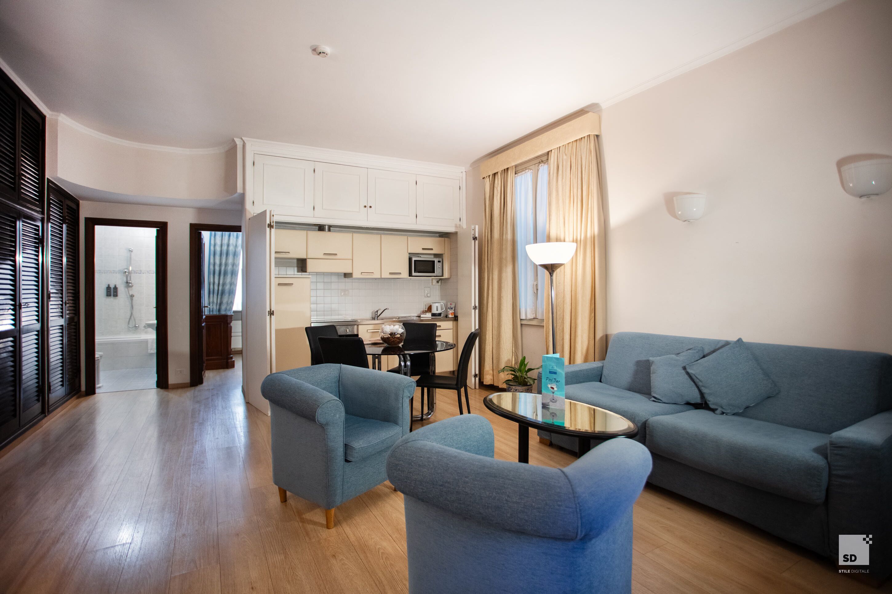Un salon moderne avec un canapé bleu, deux fauteuils, une table basse et une cuisine en arrière-plan.