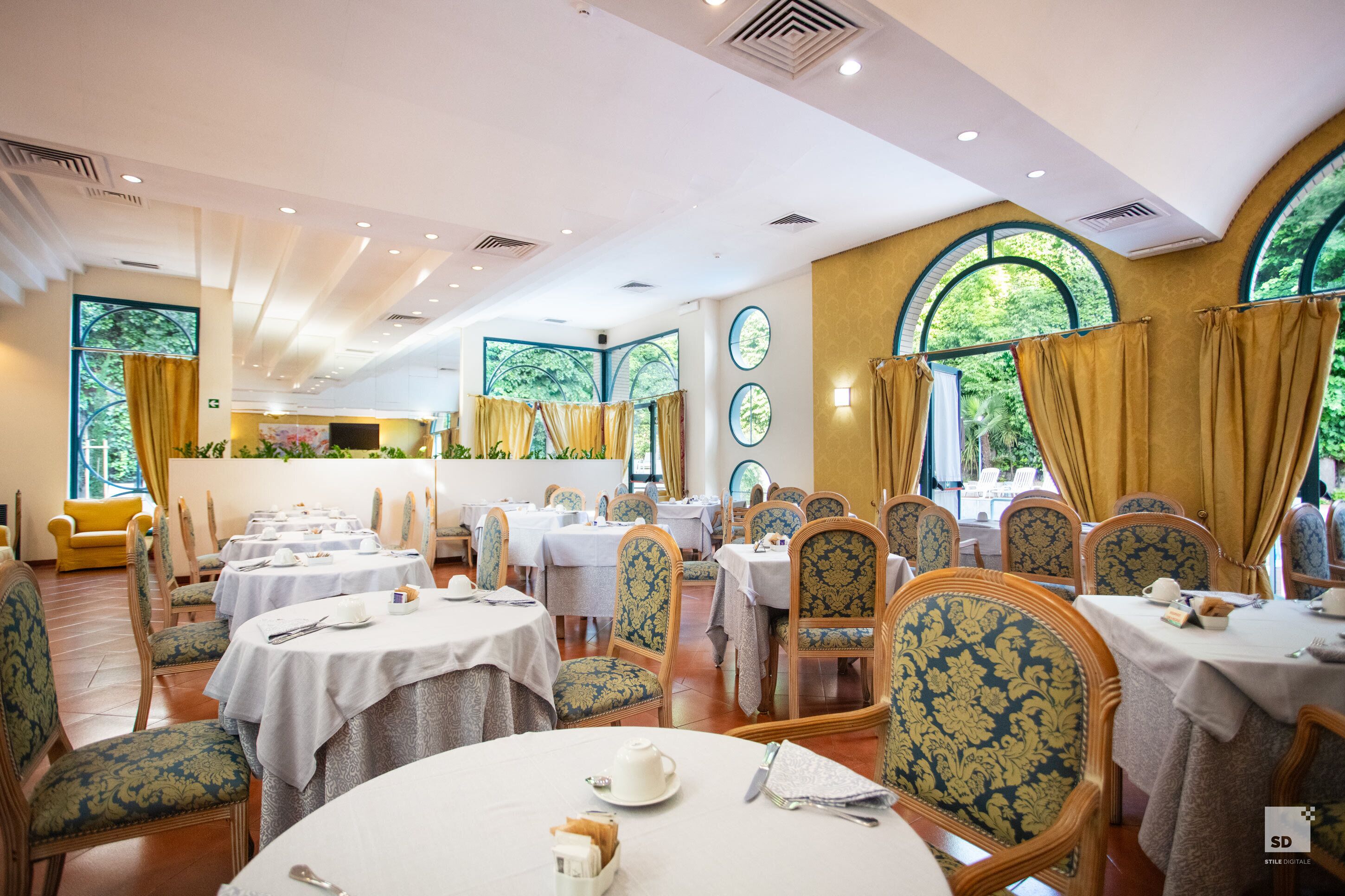Un restaurant amb taules i cadires rodones, grans finestres amb cortines, i vista a zones verdes a l'exterior.