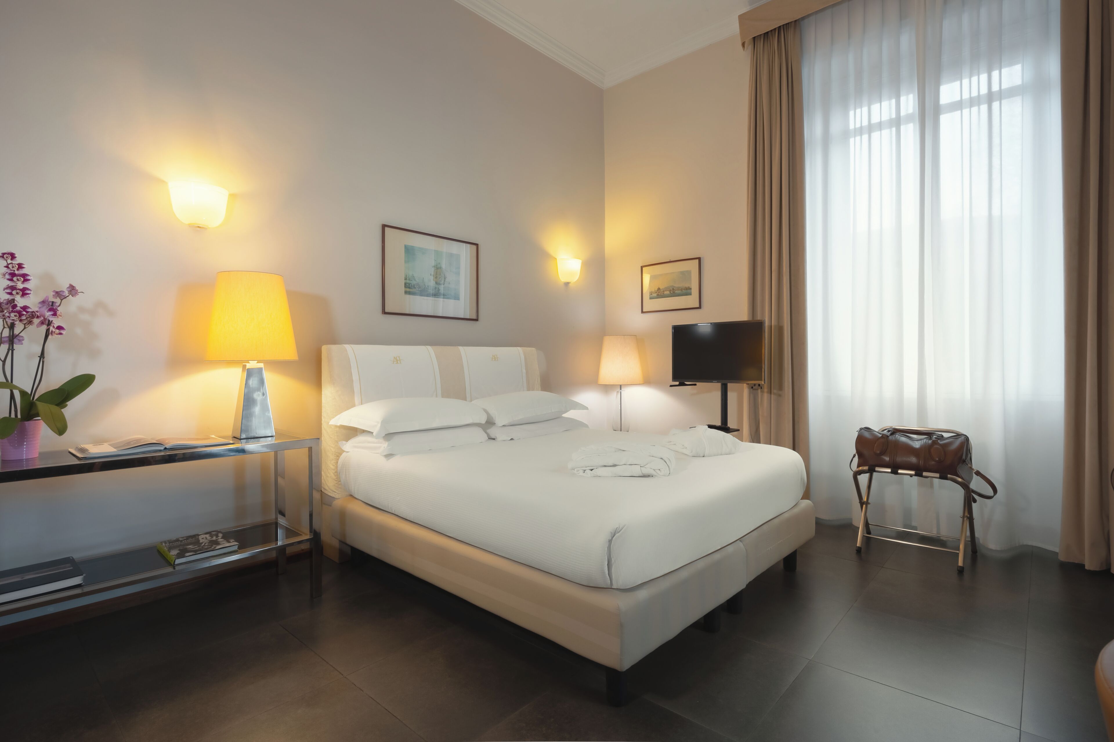 Habitación de hotel con cama, dos lámparas de noche, televisión, una mesita con lámpara, una silla y una ventana con cortinas.