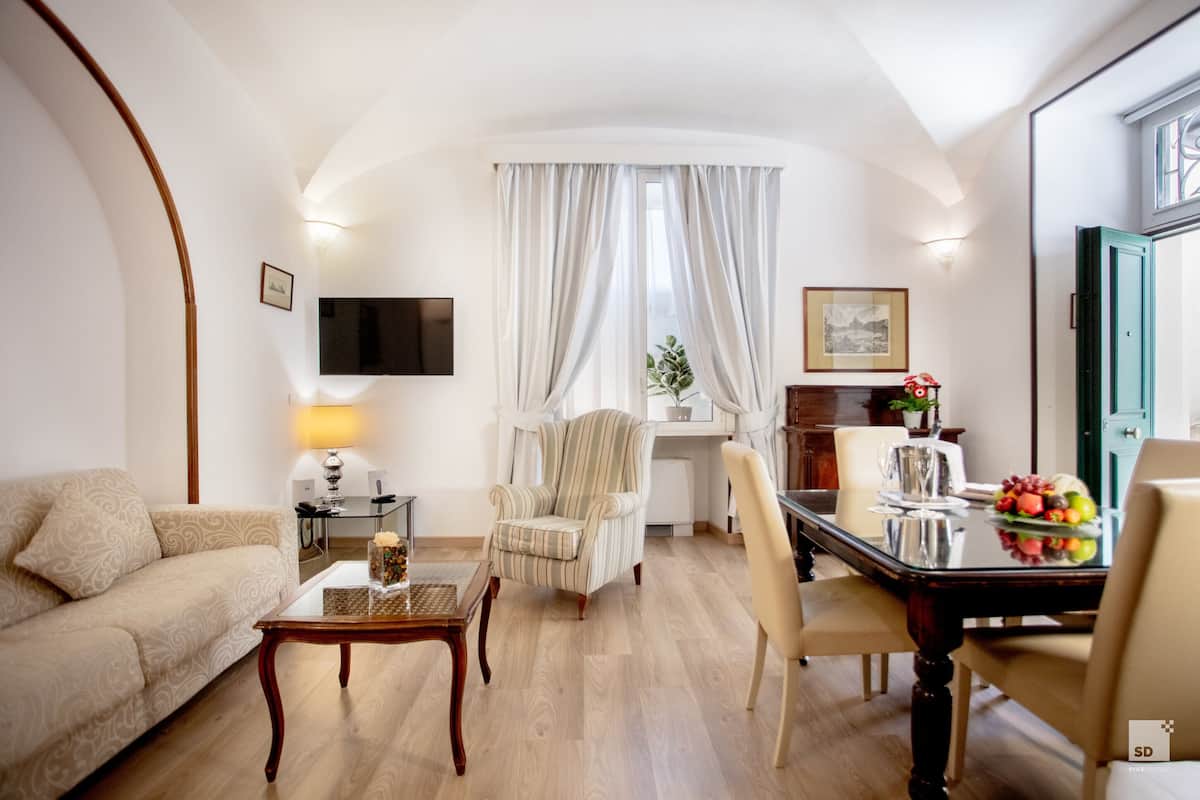 Un salon comprenant un canapé, une table basse, un fauteuil à rayures, une table à manger avec des chaises, un téléviseur fixé au mur et une fenêtre avec des rideaux.