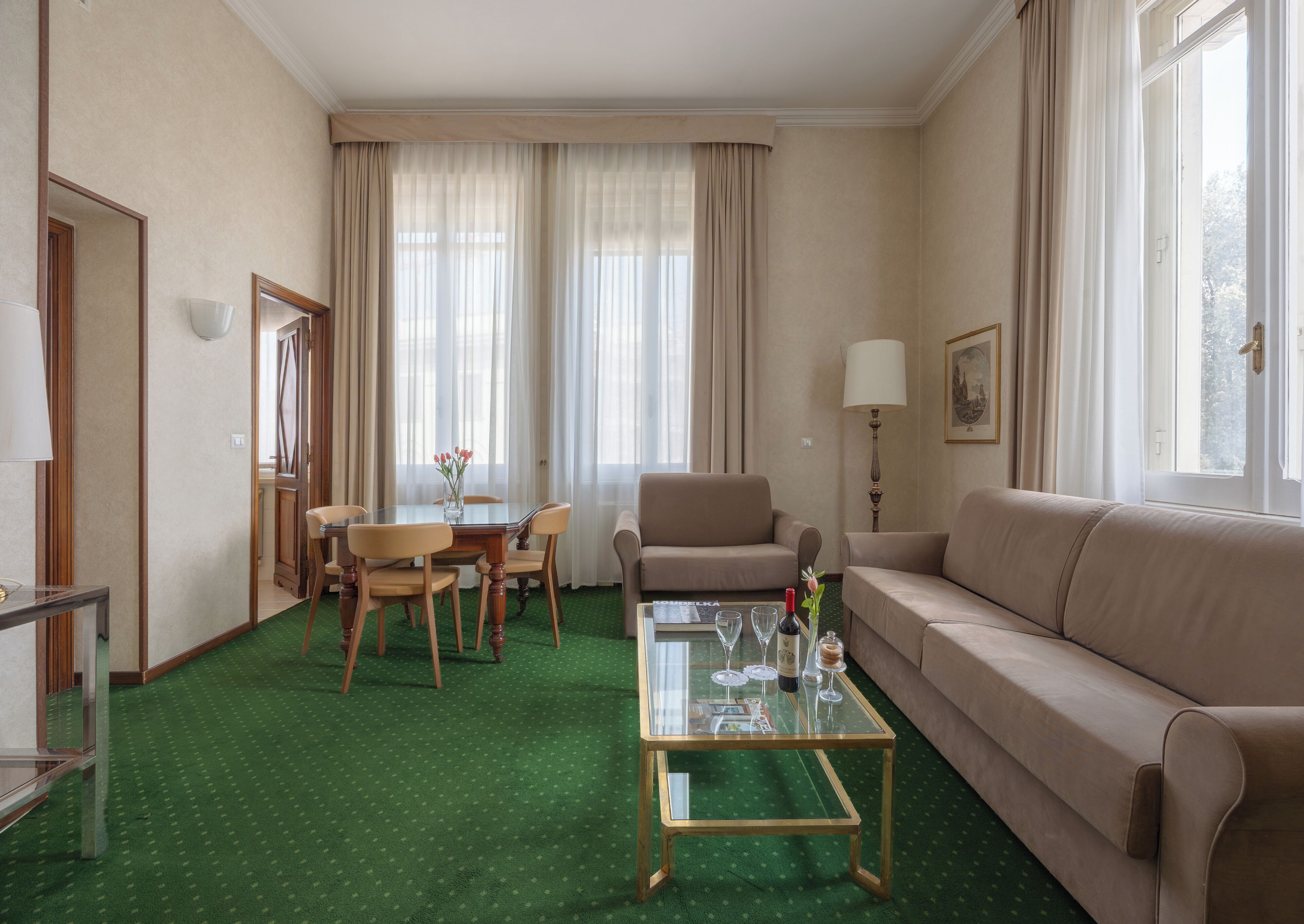 Camera d'albergo con tappeto verde, un tavolino in vetro, un divano, un tavolo da pranzo con sedie e un'ampia finestra con tende.