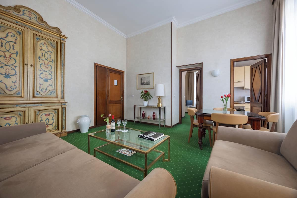 Une chambre d’hôtel avec un tapis vert, un canapé, une table basse, une table à manger avec des chaises, un buffet orné de motifs décoratifs, un miroir et un tableau encadré accroché au mur.