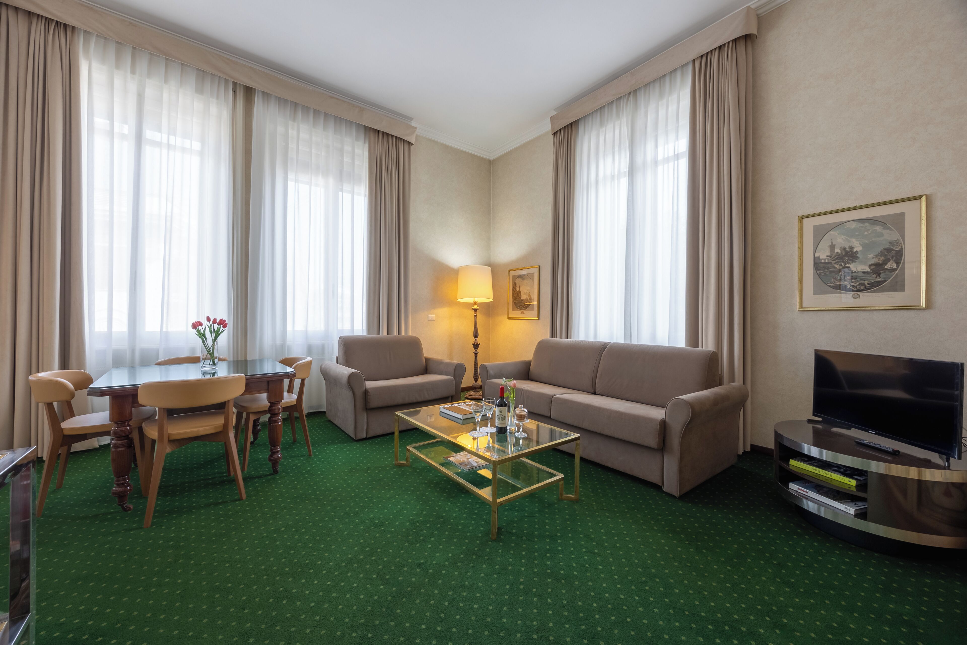 A hotel room with a green carpet, a glass coffee table, a sofa, a TV stand, and a dining table with chairs.