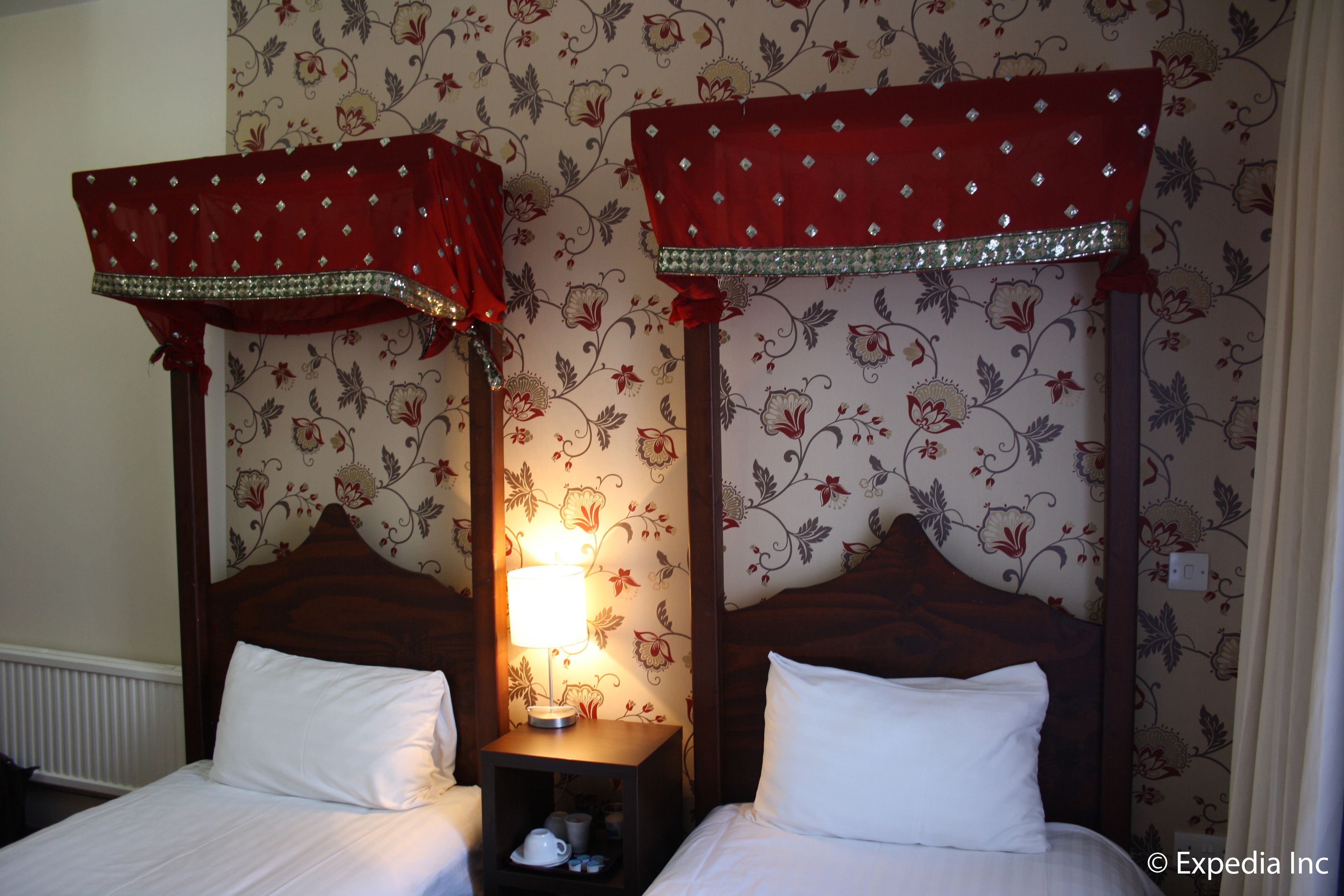 Quarto com duas camas, cada uma com cabeceira de madeira e cortinas vermelhas. Um criado-mudo com uma luminária fica entre as camas.