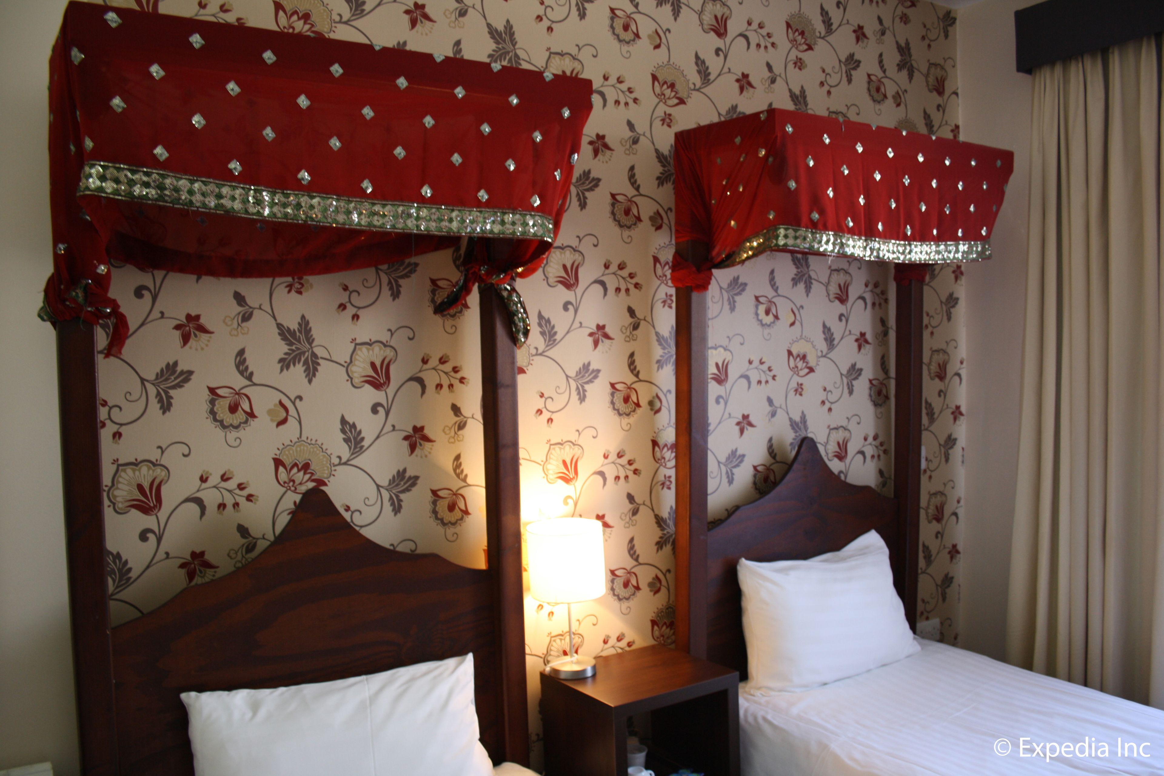 A hotel room with two beds, floral wallpaper, and red curtains with diamond patterns.