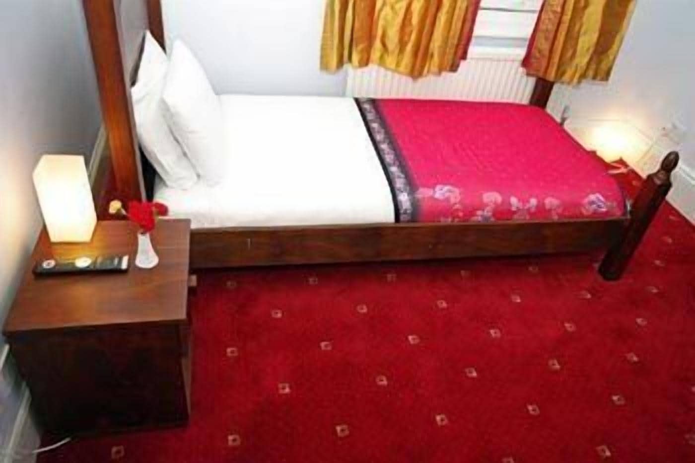 A single bed with a wooden frame, white bedding, and a red carpeted floor.