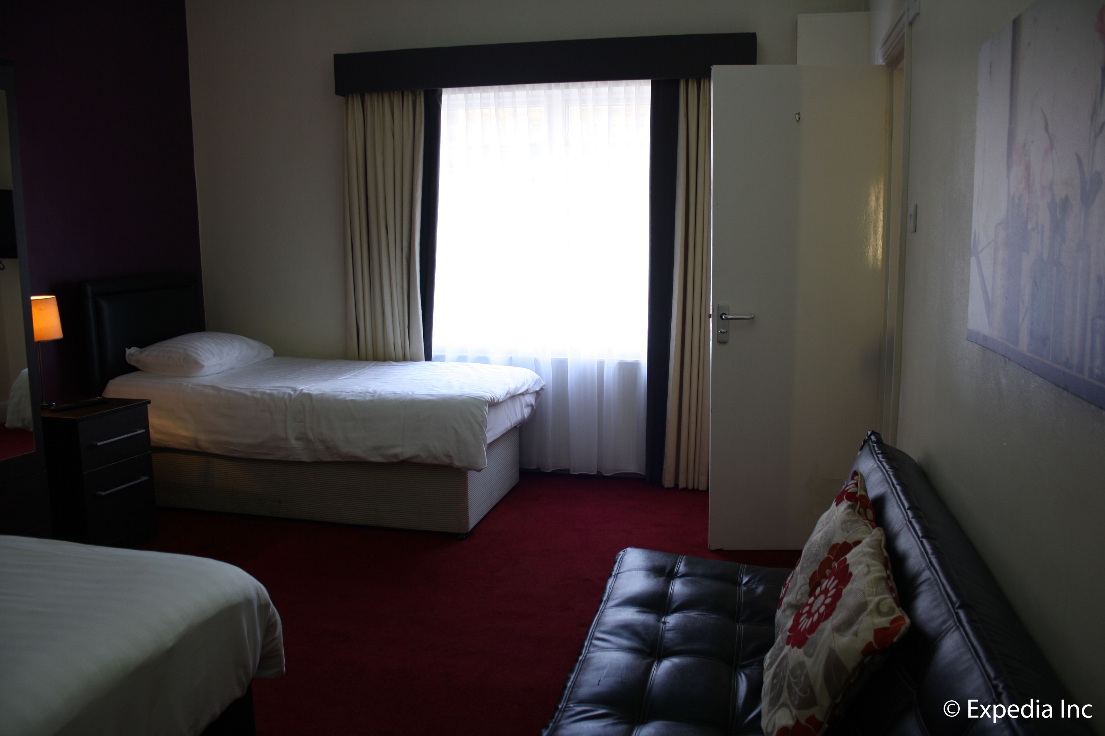 A hotel room with a bed, a bedside table, a lamp, a couch, and a window with curtains.