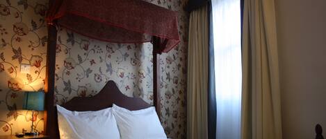 A four-poster bed with white linens, a bedside table with a lamp, and floral wallpaper.