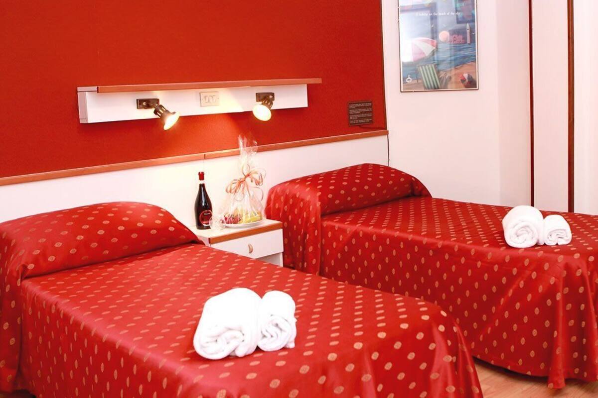A hotel room with two single beds, each with a red polka dot bedspread and white towels. There is a nightstand with a bottle and a lamp, and a suitcase on a shelf.