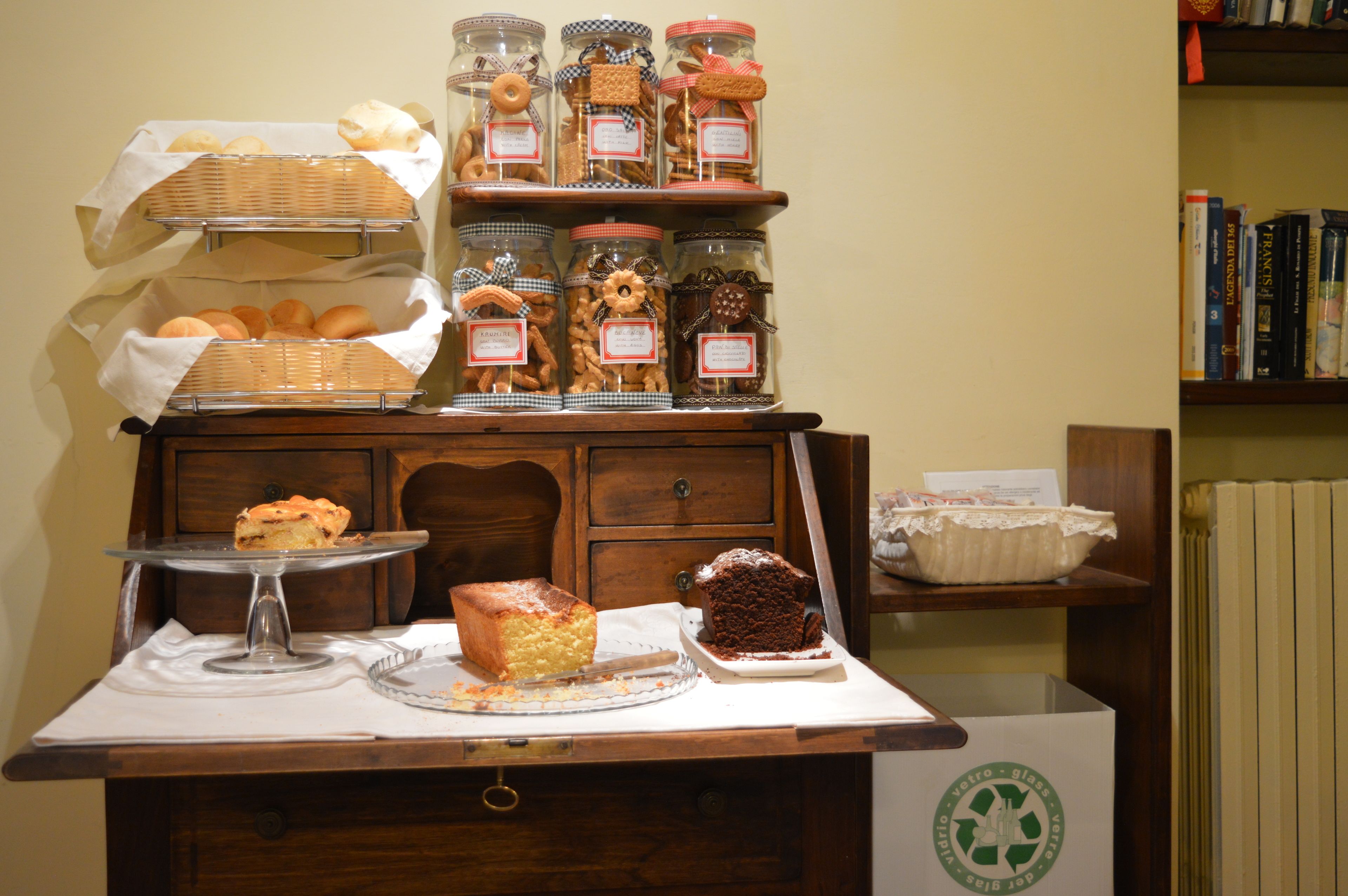 Una mesa de buffet con diversos productos horneados y pasteles, un pastel en un pedestal y un contenedor de reciclaje.