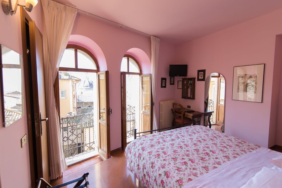 Un dormitorio con cama, escritorio, silla y televisor. Hay dos ventanas con cortinas, y se ve un balcón a través de una ventana.