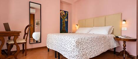 Un dormitorio con cama, escritorio con laptop, silla, espejo y un cuadro en la pared.