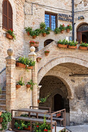 A stone building with a sign reading 'HOTEL PALLOTTA.' There are multiple flower pots with plants on the facade, and a wooden bench area in the foreground. - Hotel Pallotta Assisi (Assisi)