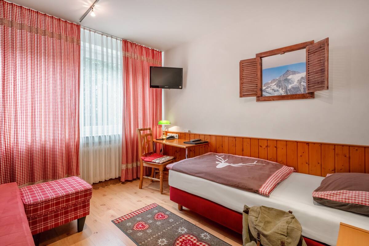 Une chambre à coucher comprenant un lit, un bureau, une chaise, un téléviseur et une fenêtre avec des rideaux.