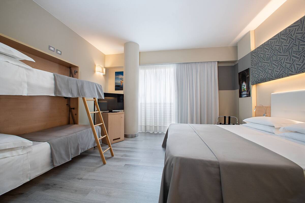 Een hotelkamer met een stapelbed, een bureau met stoel, een ladder en een groot raam met gordijnen.